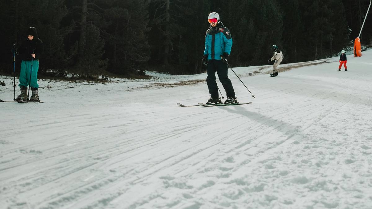 Osoba na nartach zjeżdżająca po ośnieżonym stoku narciarskim, w tle inne osoby na nartach i drzewa.
