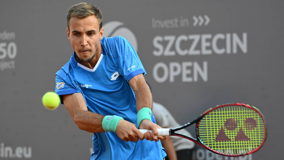 Mężczyzna grający w tenisa, uderzający piłkę rakietą. Nosi niebieską koszulkę i opaski na nadgarstkach.