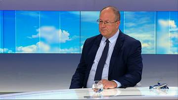 Nowy prezes PKOl siedzi przy białym stole w studio telewizyjnym na tle niebieskiego ekranu