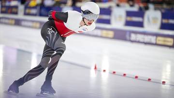 Łyżwiarz szybki w białym i czerwonym kombinezonie pochylony na torze lodowym.