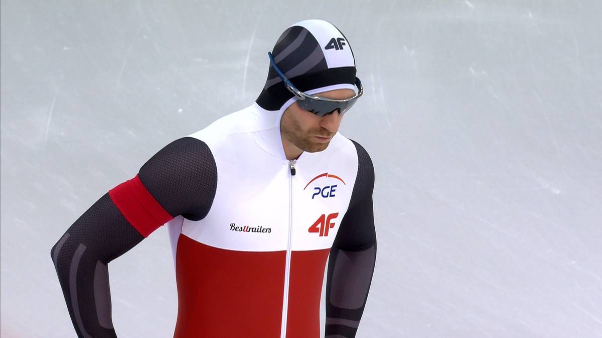 Mężczyzna w stroju łyżwiarza szybkiego, w białej czapce z paskami, goglach i stroju sportowym w kolorach białym, czerwonym i czarnym.