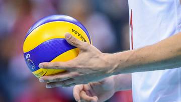 Siatkarz w białej koszulce z czerwonymi rękawami i czerwonych spodenkach trzyma piłkę siatkową w dłoniach.