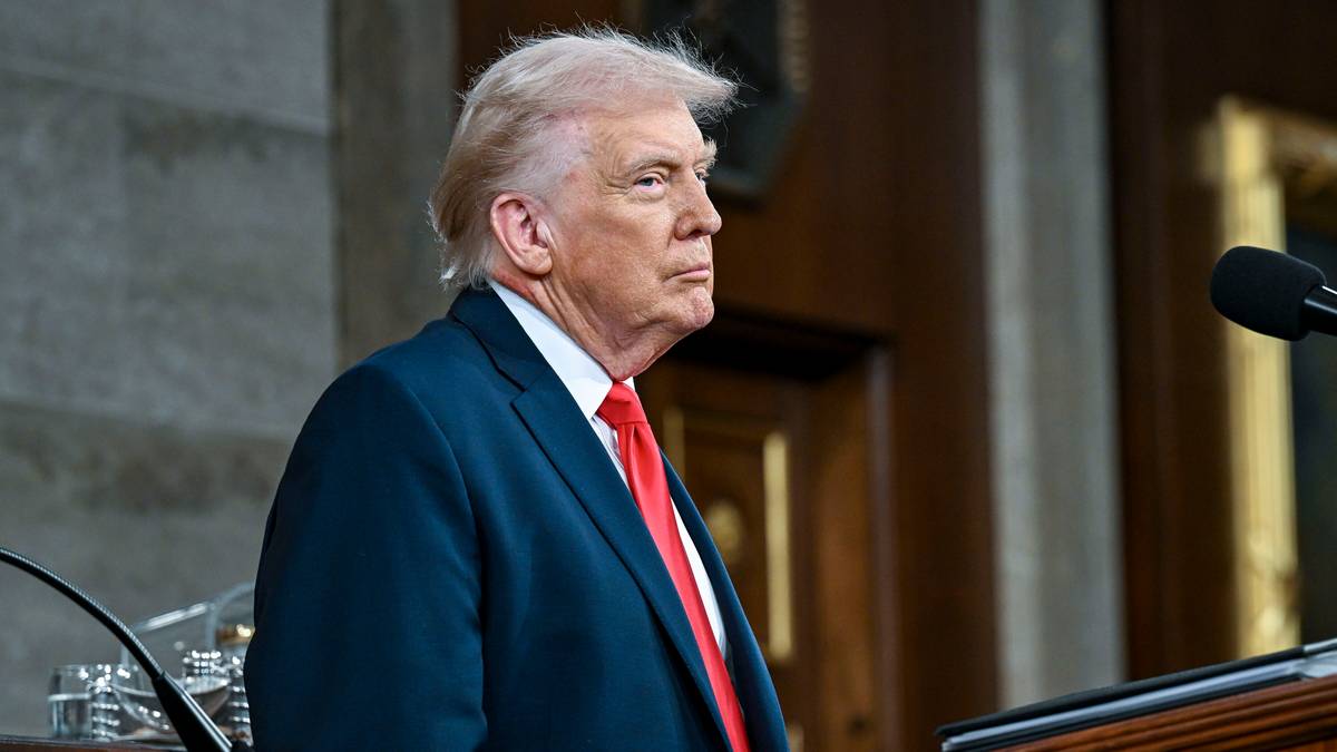 Donald Trump w granatowym garniturze i czerwonym krawacie stoi w pomieszczeniu, patrząc w bok.