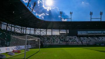 Widok na pusty stadion piłkarski z murawą, bramką i trybunami pod błękitnym niebem z promieniami słońca przebijającymi się przez chmury.