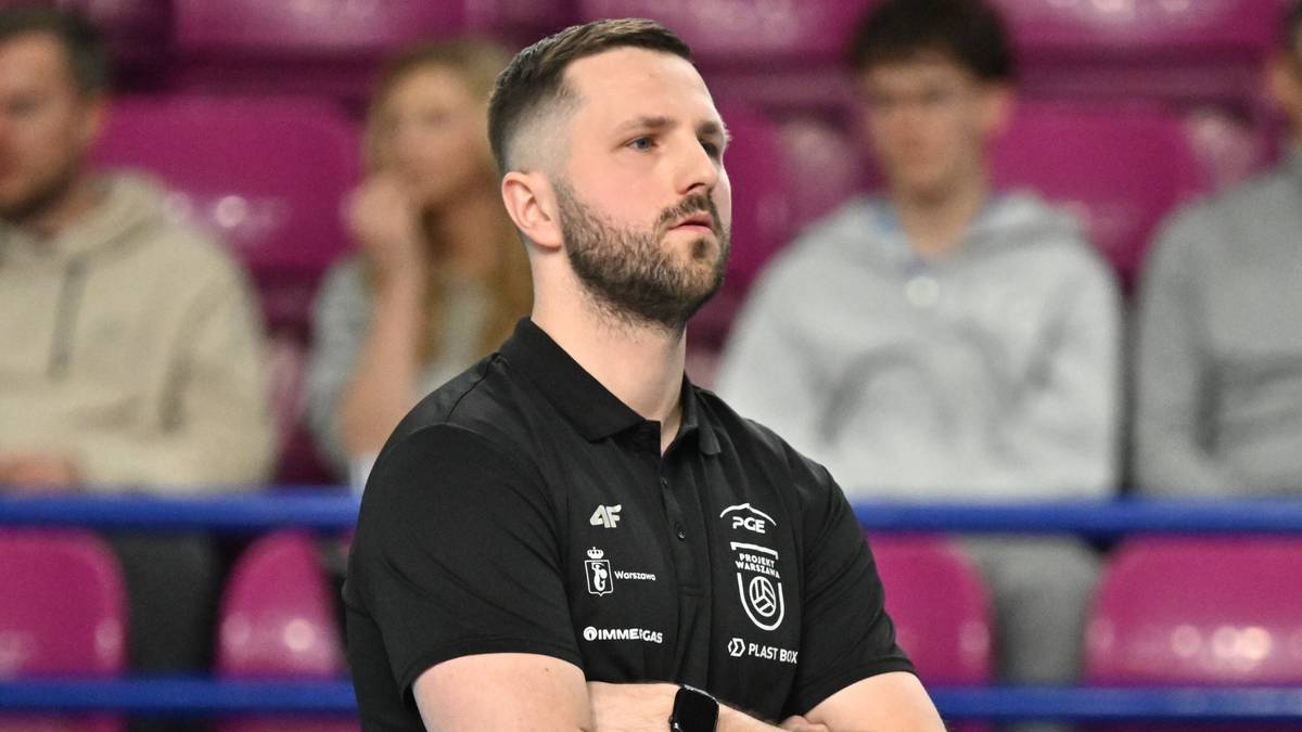 Trener PGE Projekt Warszawa stoi na boisku z rękami skrzyżowanymi na piersi, w tle widzowie na trybunach.