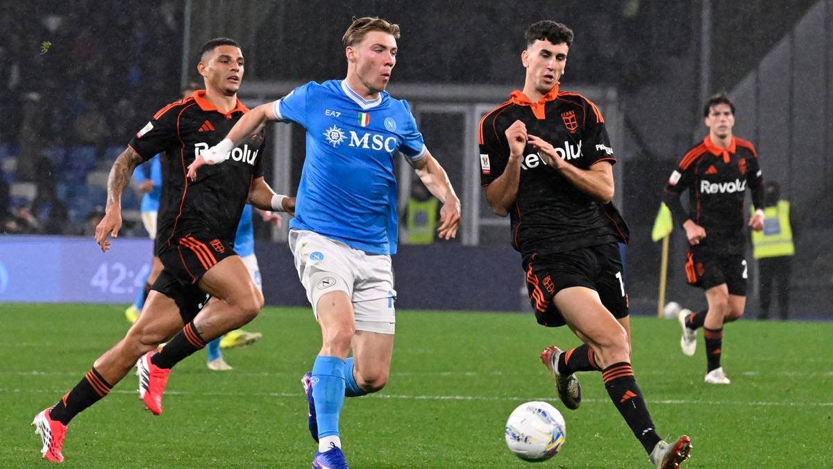 Trzech piłkarzy w niebieskich i czarno-pomarańczowych strojach ściga się za piłką na zielonej murawie stadionu piłkarskiego.