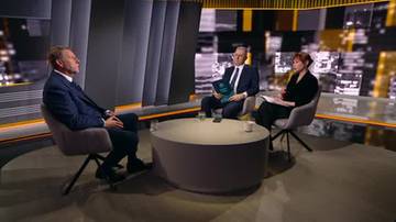 Trzy osoby siedzą przy okrągłym stole w studiu telewizyjnym: dw&oacute;ch mężczyzn w garniturach i jedna kobieta.