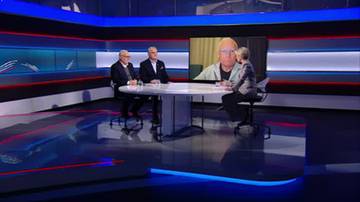Trzech mężczyzn siedzi przy owalnym stole w studiu telewizyjnym. Na dużym ekranie w tle widoczny jest czwarty mężczyzna.