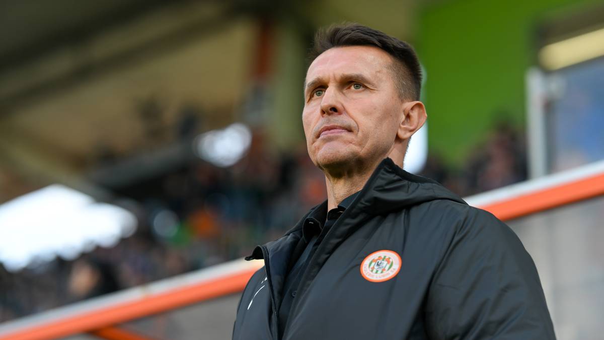 Mężczyzna w czarnej kurtce z naszywką klubu sportowego na tle trybun stadionu.