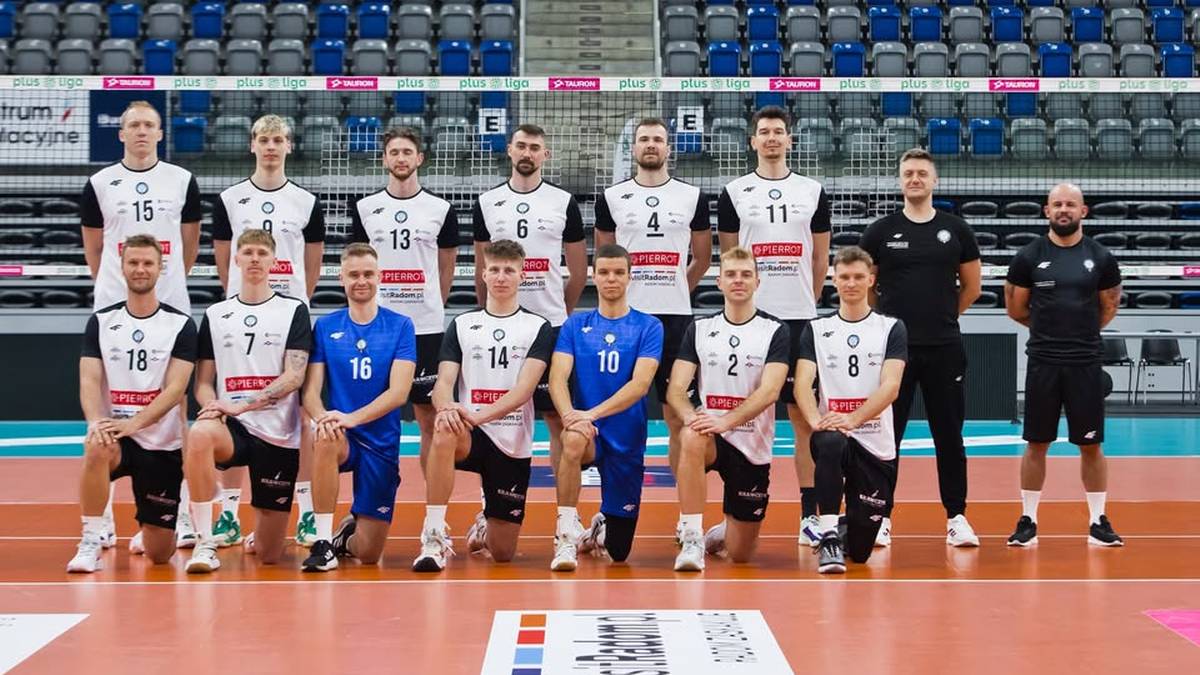 Drużyna siatkarska w białych i czarnych strojach pozuje do zdjęcia na tle trybun.