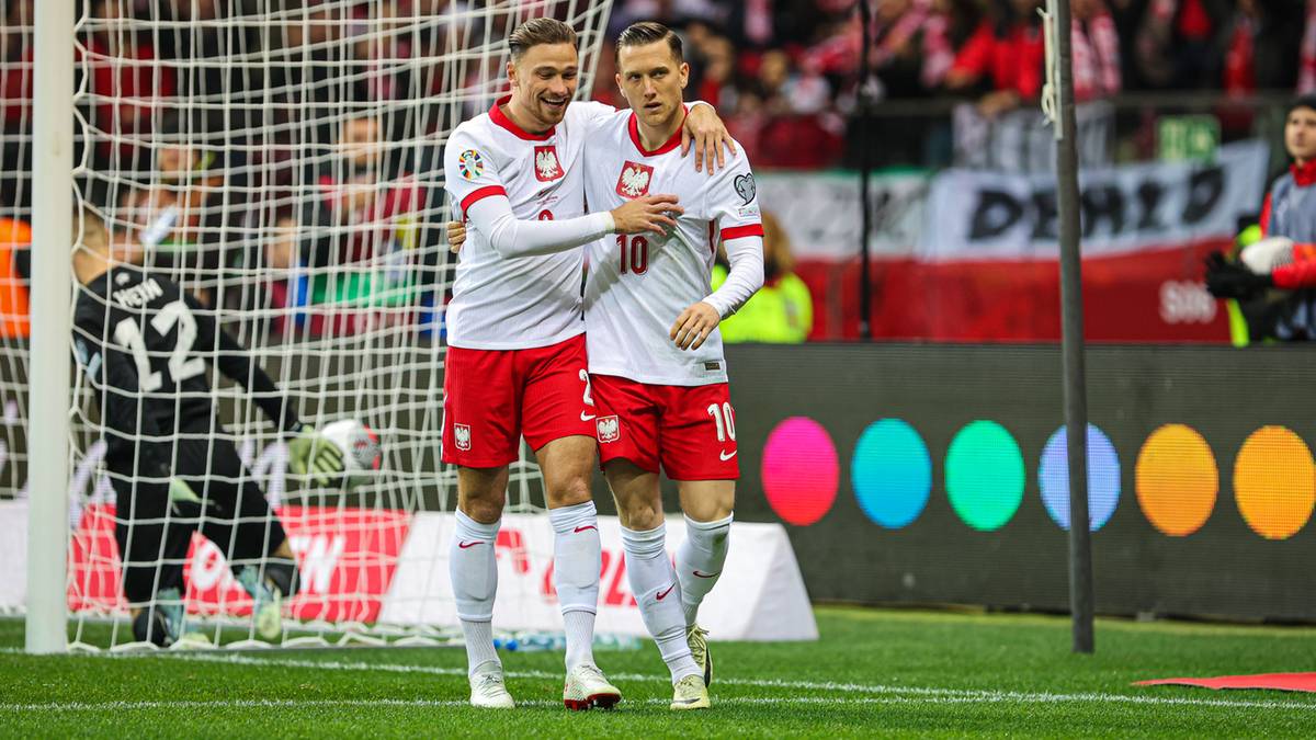 Dwóch piłkarzy reprezentacji Polski w białych strojach świętuje przy bramce, w tle kibice na stadionie.