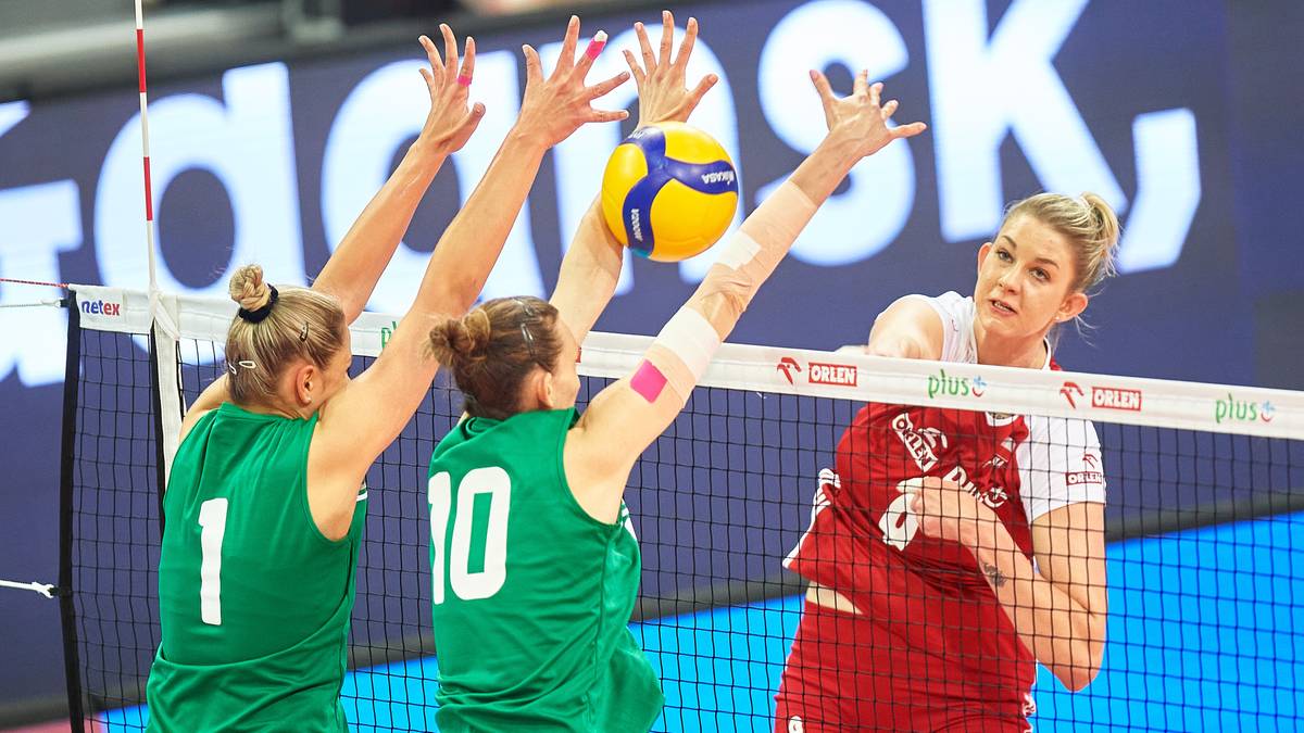 Trzy siatkarki w akcji nad siatką, dwie w zielonych strojach próbują zablokować atak zawodniczki w czerwonym stroju.