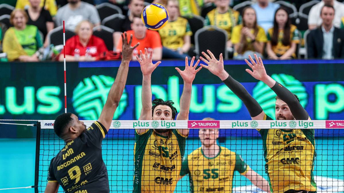 Siatkarze podczas meczu blokują piłkę nad siatką. Gracz w czarnej koszulce z numerem 9 skacze do ataku, podczas gdy gracze w żółtych koszulkach z numerami 15 i 55 próbują go zablokować.
