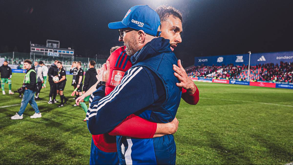 Dw&oacute;ch mężczyzn w strojach sportowych przytula się na tle stadionu piłkarskiego.
