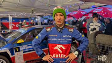 Szymon Gospodarczyk w czapce z pomponem, ubrany w kombinezon rajdowy z logo Orlen i Skoda, stoi przed samochodem rajdowym.
