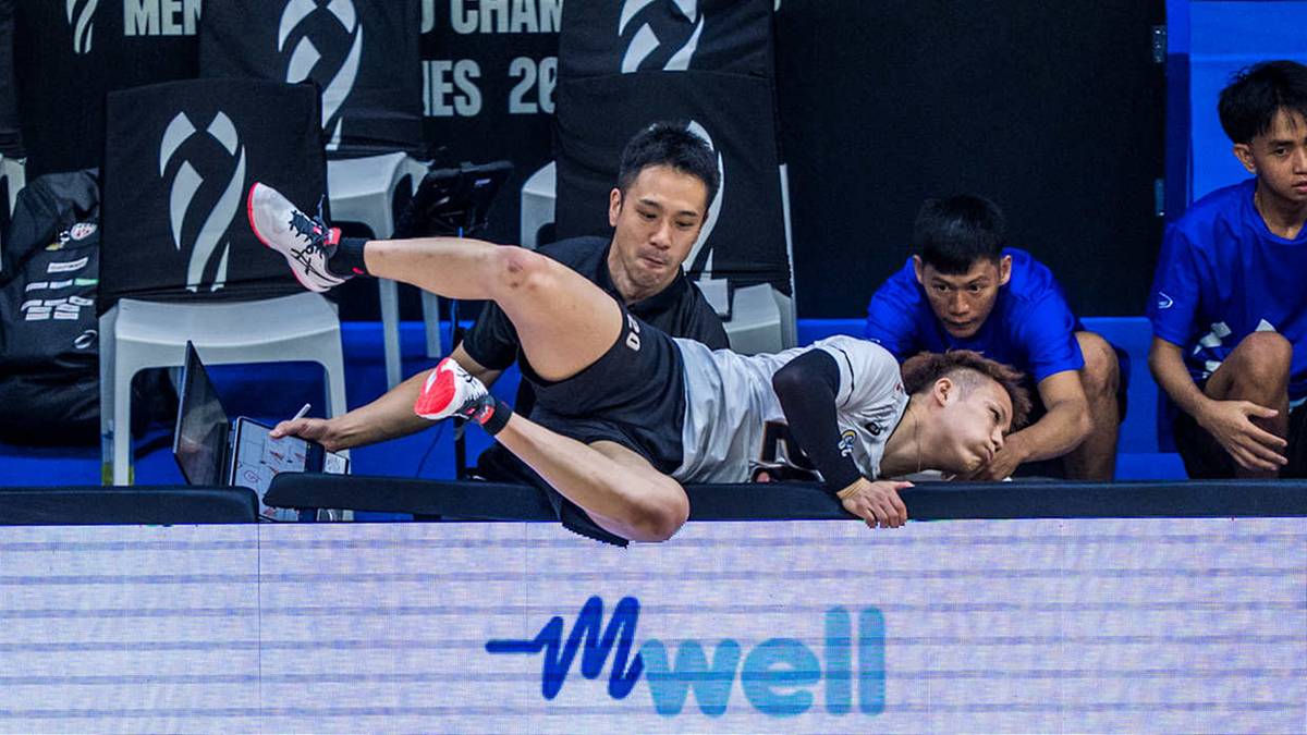 Siatkarz w czarnym stroju upada na parkiet, próbując obronić piłkę. Obok niego leży inny zawodnik, a za nimi widoczni są inni członkowie drużyny.