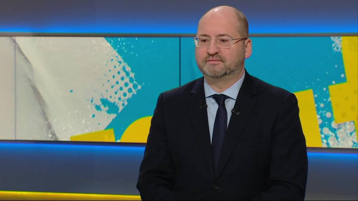 Mężczyzna w ciemnym garniturze i niebieskiej koszuli z krawatem, w okularach, siedzi w studiu telewizyjnym na tle ekranu z abstrakcyjnym wzorem w kolorach białym, niebieskim i żółtym.