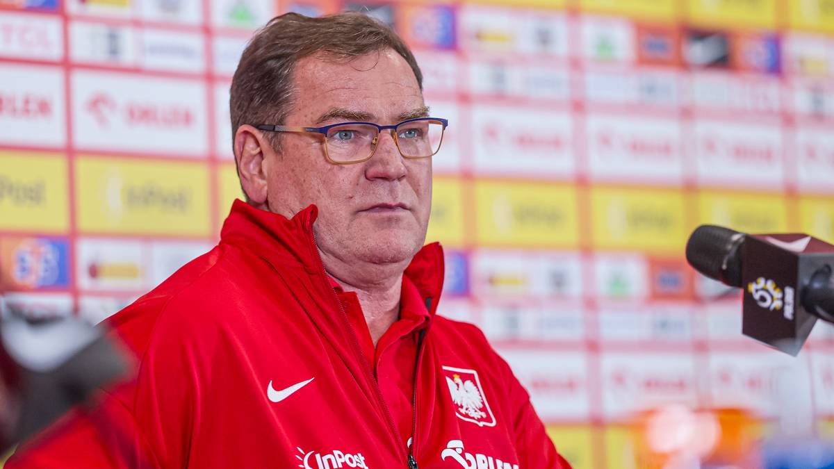 Portret Jana Urbana, trenera piłkarskiego, noszącego okulary i czerwoną bluzę z orłem na piersi. Stoi przed ścianą z logo.