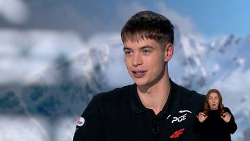 Młody mężczyzna w czarnej koszulce sportowej z logo PGE i 4F, mówiący. W tle widoczna jest osoba tłumacząca język migowy.