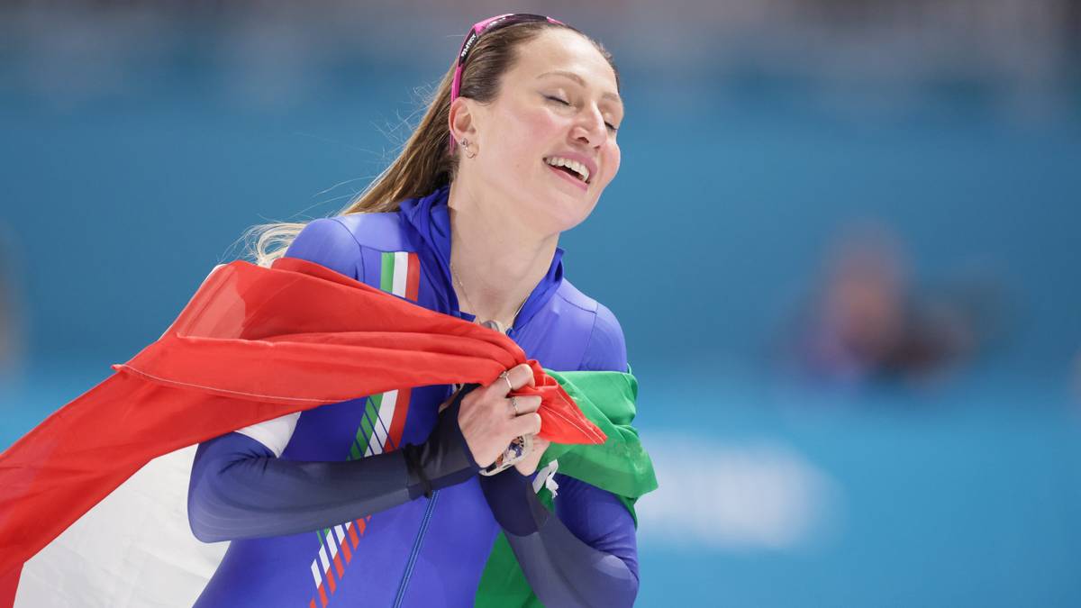 Portret kobiety w niebieskim stroju sportowym, trzymającej we włoską flagę, z uśmiechem na twarzy i zamkniętymi oczami.