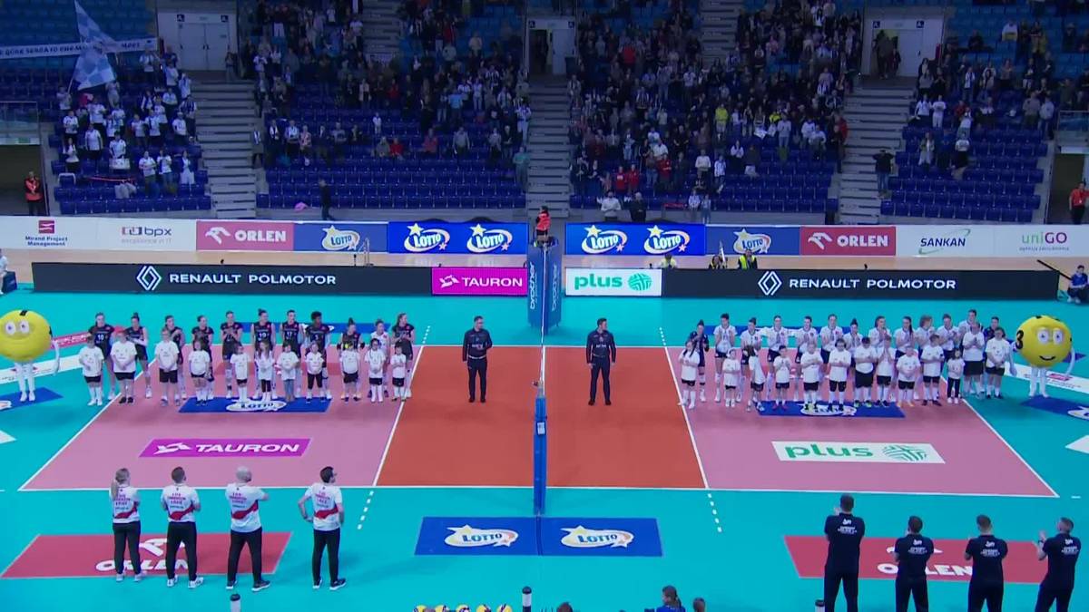 Dwie drużyny siatkarzy stojące naprzeciwko siebie na boisku w hali sportowej, z trybunami wypełnionymi widzami w tle.