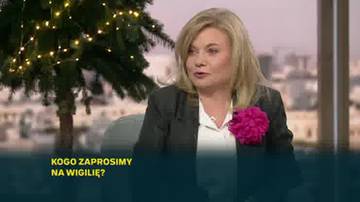 Kobieta w ciemnej marynarce i białej bluzce z ozdobnym przypinką w kształcie pomponu, siedząca przed choinką ozdobioną lampkami. Na dole ekranu widnieje napis "KOGO ZAPROSIMY NA WIGIILIĘ?".