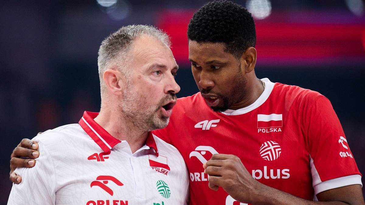 Dwóch mężczyzn w strojach sportowych, prawdopodobnie siatkarze lub trener i zawodnik, rozmawiają ze sobą na ciemnym tle.