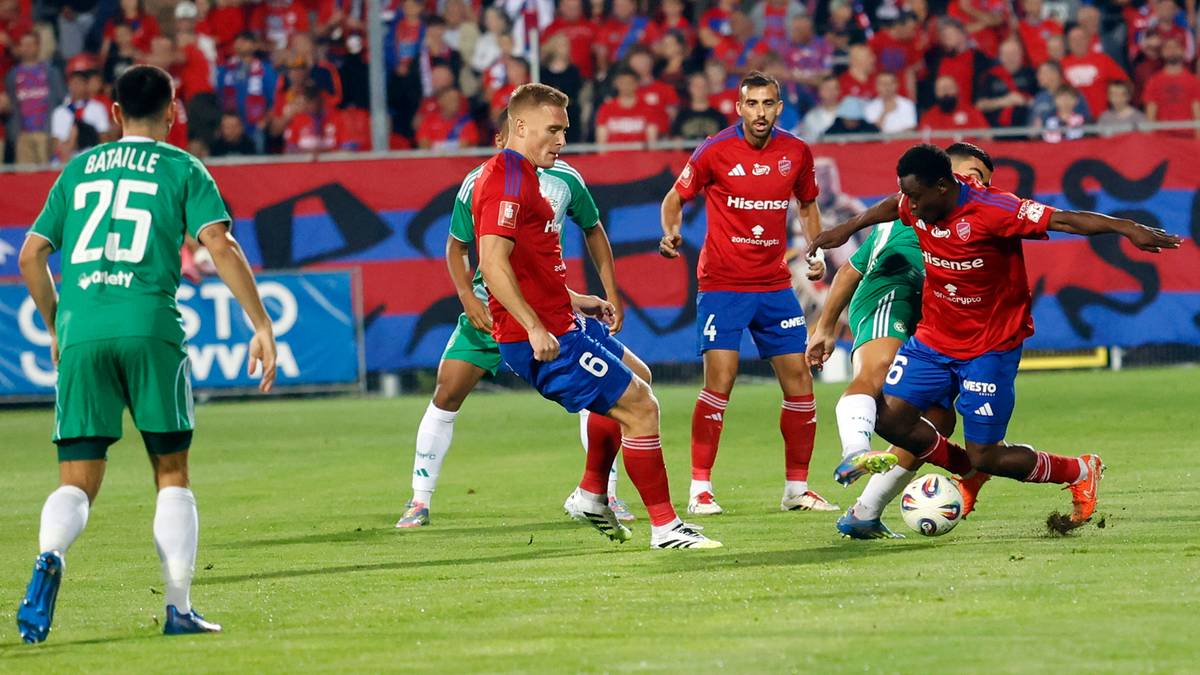 Piłkarze w czerwonych i zielonych strojach walczą o piłkę na boisku piłkarskim, z publicznością w tle.