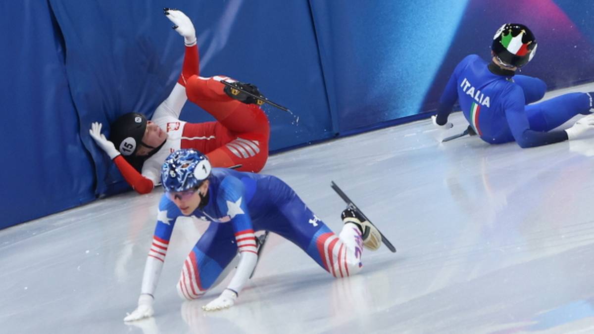 Trzy łyżwiarki na lodowisku short track po upadku jednej z nich, która leży na lodzie.
