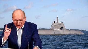 Mężczyzna w okularach i garniturze przemawia do mikrofon&oacute;w, na tle widoczny okręt podwodny.