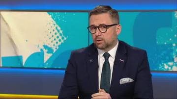 Mężczyzna w okularach i ciemnym garniturze z zielonym krawatem siedzi przed ekranem z kolorowym wzorem.