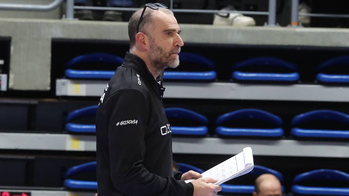 Trener siatk&oacute;wki w czarnym stroju trzymający notatnik, w tle trybuny hali sportowej.