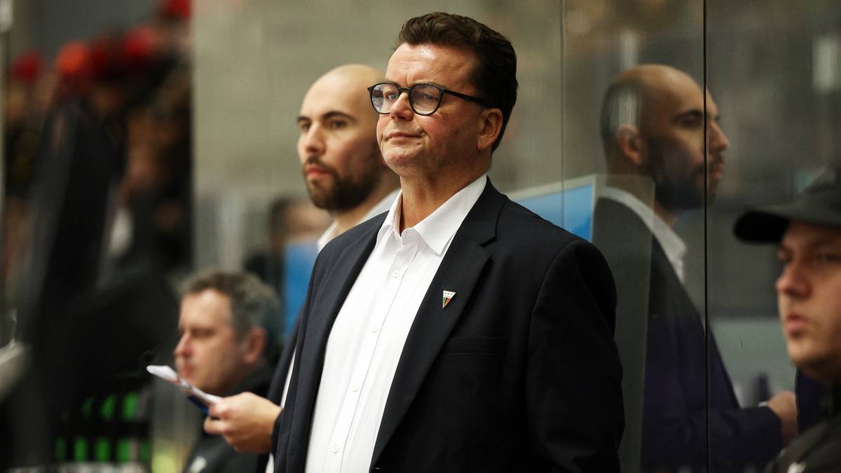 Mężczyzna w okularach, ubrany w białą koszulę i marynarkę, stoi na tle lodowiska, patrząc w bok.