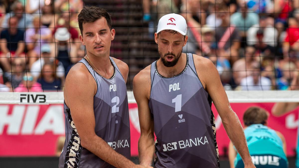 Dwóch polskich siatkarzy plażowych w szarych koszulkach z numerami 1 i 2 na piersiach, patrzących na siebie z powagą.