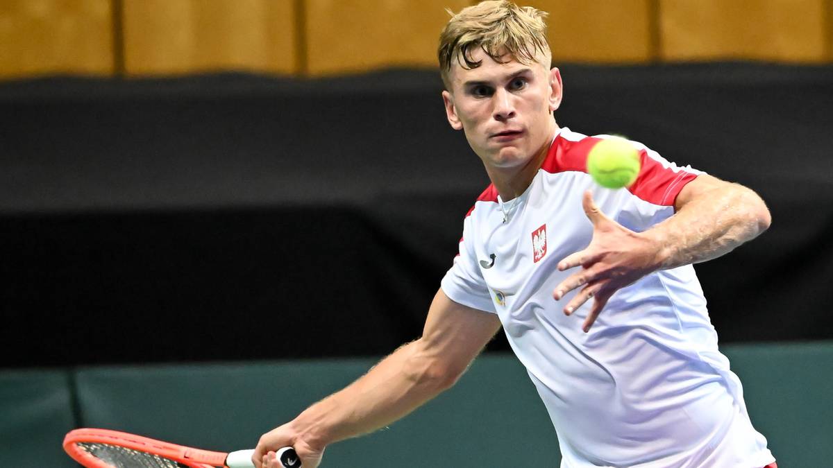 Młody mężczyzna w białej koszulce z godłem Polski, wykonuje uderzenie tenisowe rakietą.