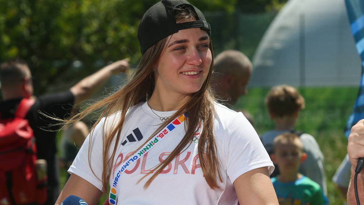 Młoda kobieta w czapce i białej koszulce z napisem "POLSKA" i ozdobnym pasem, uśmiecha się patrząc w bok. Tło jest rozmyte i przedstawia innych ludzi oraz elementy budowli.