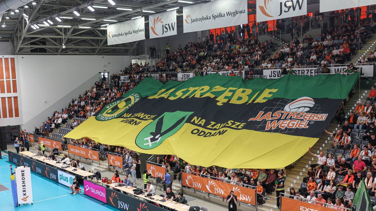 Kibice na trybunach hali sportowej trzymają ogromną czarno-zielono-ż&oacute;łtą flagę z napisem JASTRZĘBIE.