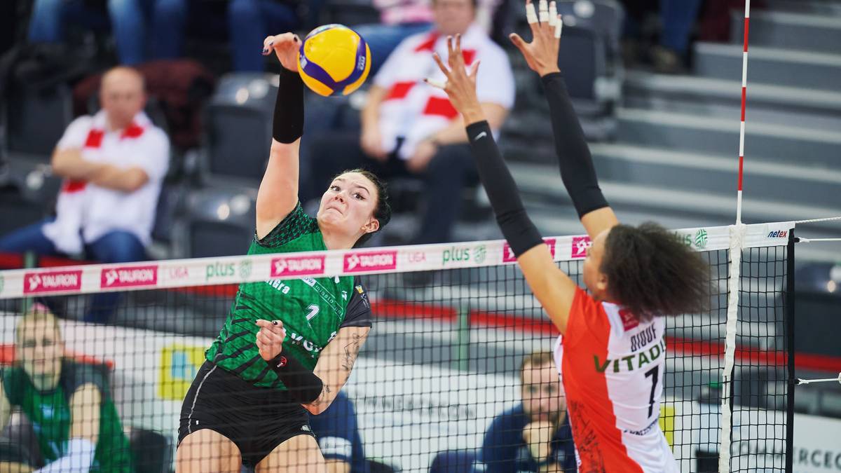 Siatkarka w zielonej koszulce wykonuje zagrywkę, blokując ją przeciwniczka w czerwonej koszulce.