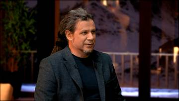 Łukasz Szeliga, mężczyzna z siwymi włosami i brodą, ubrany w ciemną marynarkę, udziela wywiadu.