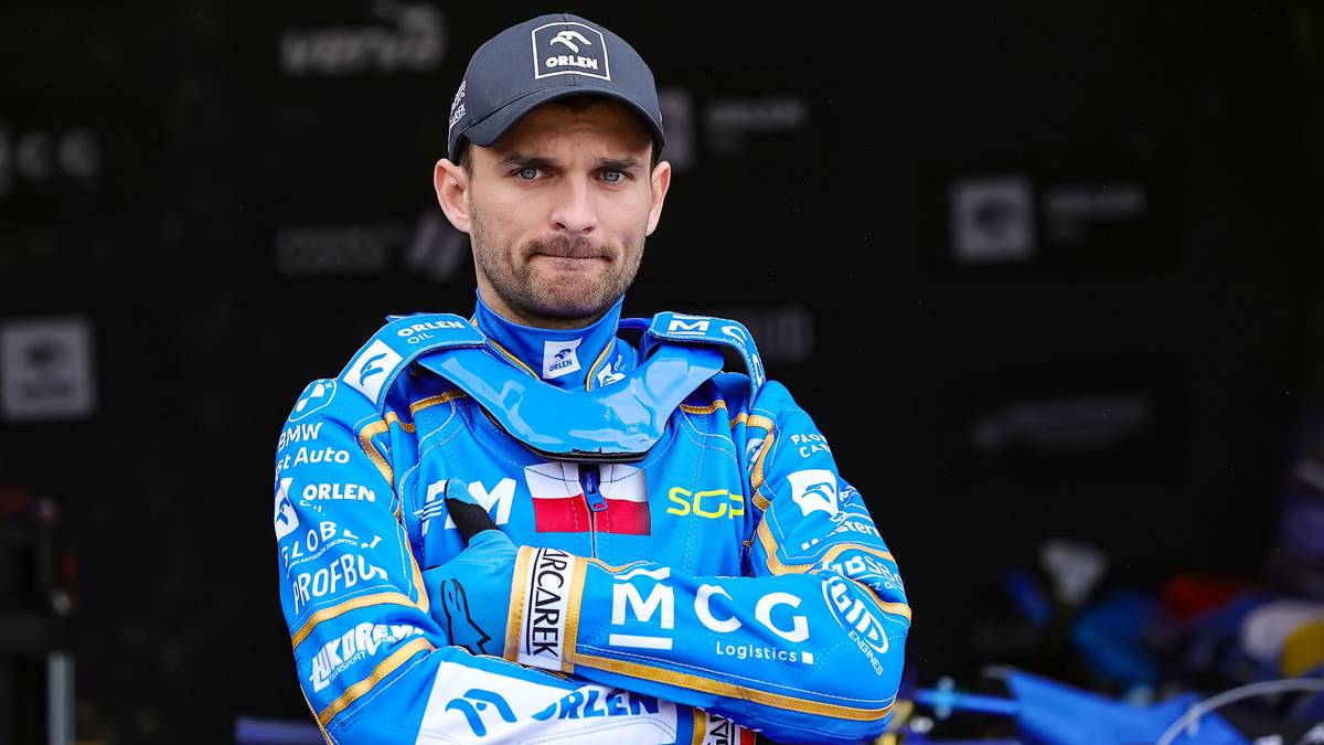 Żużlowiec w niebieskim kevlarze i czapce z daszkiem, z godłem Polski na piersi.