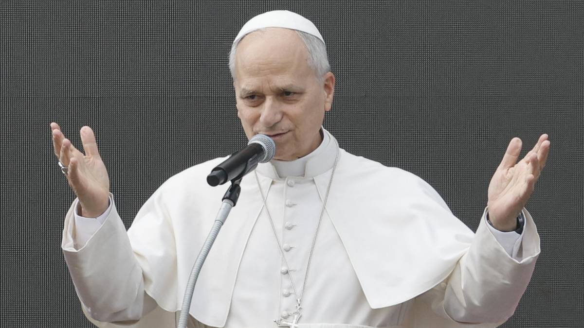 Papież w białej sutannie i piusce przemawia do mikrofonu, unosząc ręce w geście błogosławieństwa.