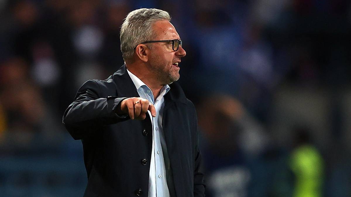 Jerzy Brzęczek, w okularach i ciemnej marynarce, stoi z wyciągniętą ręką w kierunku czegoś poza kadrem, patrząc w bok.