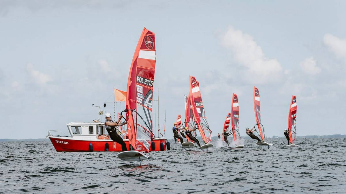 Grupa osób uprawiających windsurfing na wodzie. W tle widać czerwoną łódź z białym nadbudówką.