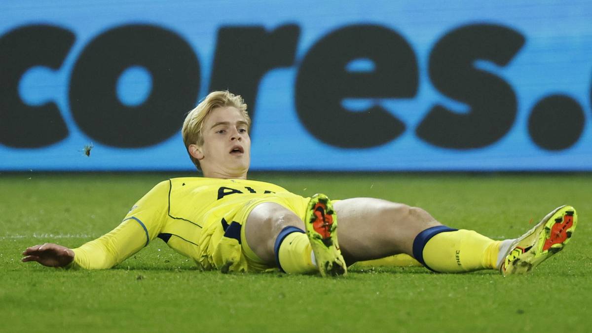 Młody piłkarz leży na murawie boiska piłkarskiego, ubrany w żółty strój z niebieskimi elementami i numerem na plecach, z wyrazem bólu na twarzy.