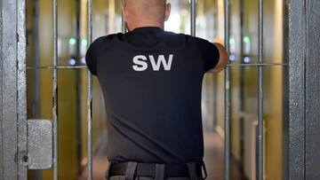 Funkcjonariusz Służby Więziennej w czarnej koszulce z napisem SW patrzący przez metalowe kraty.