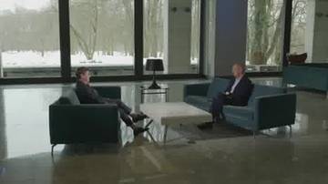 Dw&oacute;ch mężczyzn siedzi naprzeciwko siebie na ciemnych kanapach w jasnym pomieszczeniu z dużym oknem zimą.