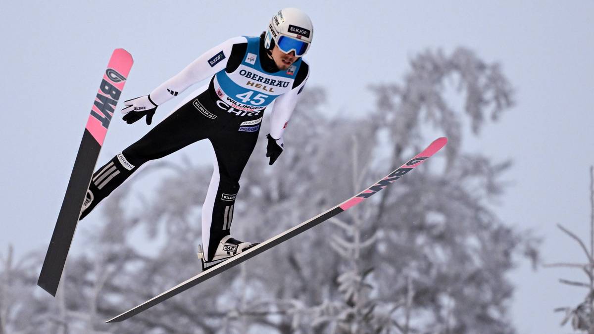 Skoczek narciarski w locie, ubrany w biały kombinezon z czarnymi elementami i numerem 45, w białym kasku i goglach.