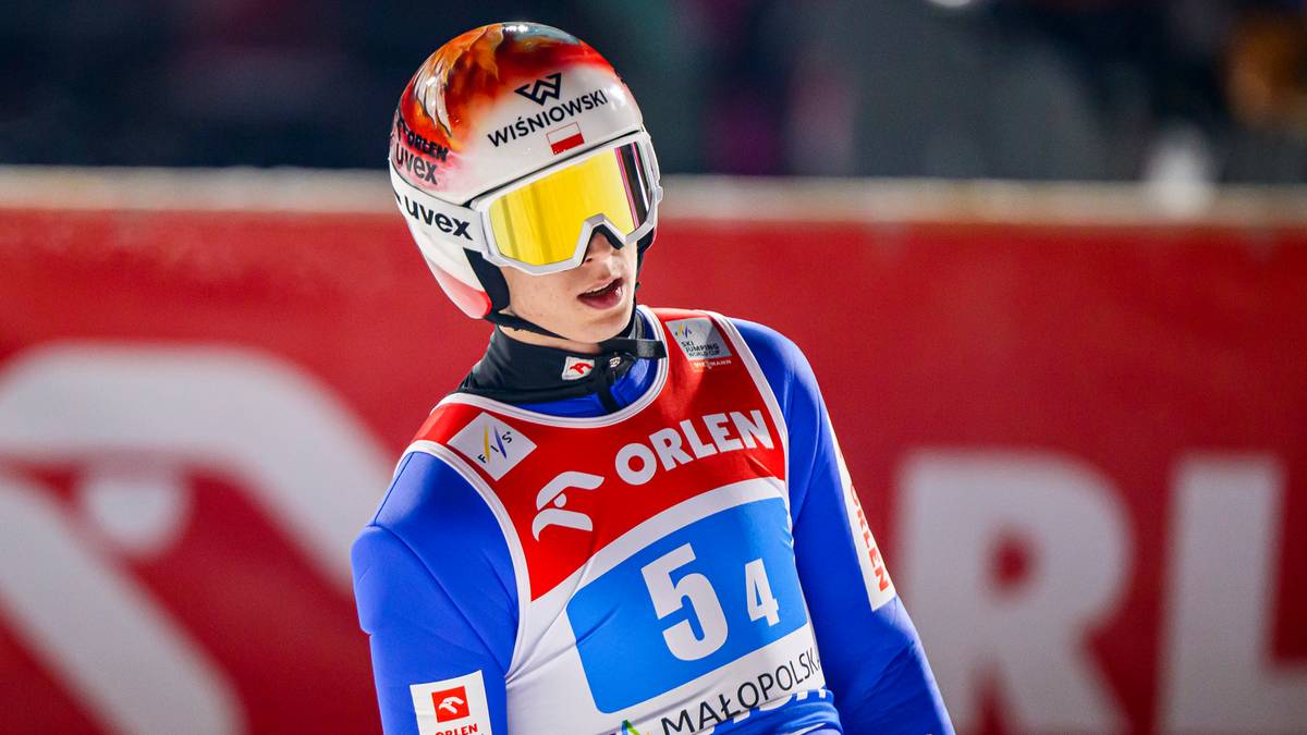 Skoczek narciarski w niebiesko-biało-czerwonym stroju i kasku z ż&oacute;łtymi goglami, z numerem 54 na piersi.