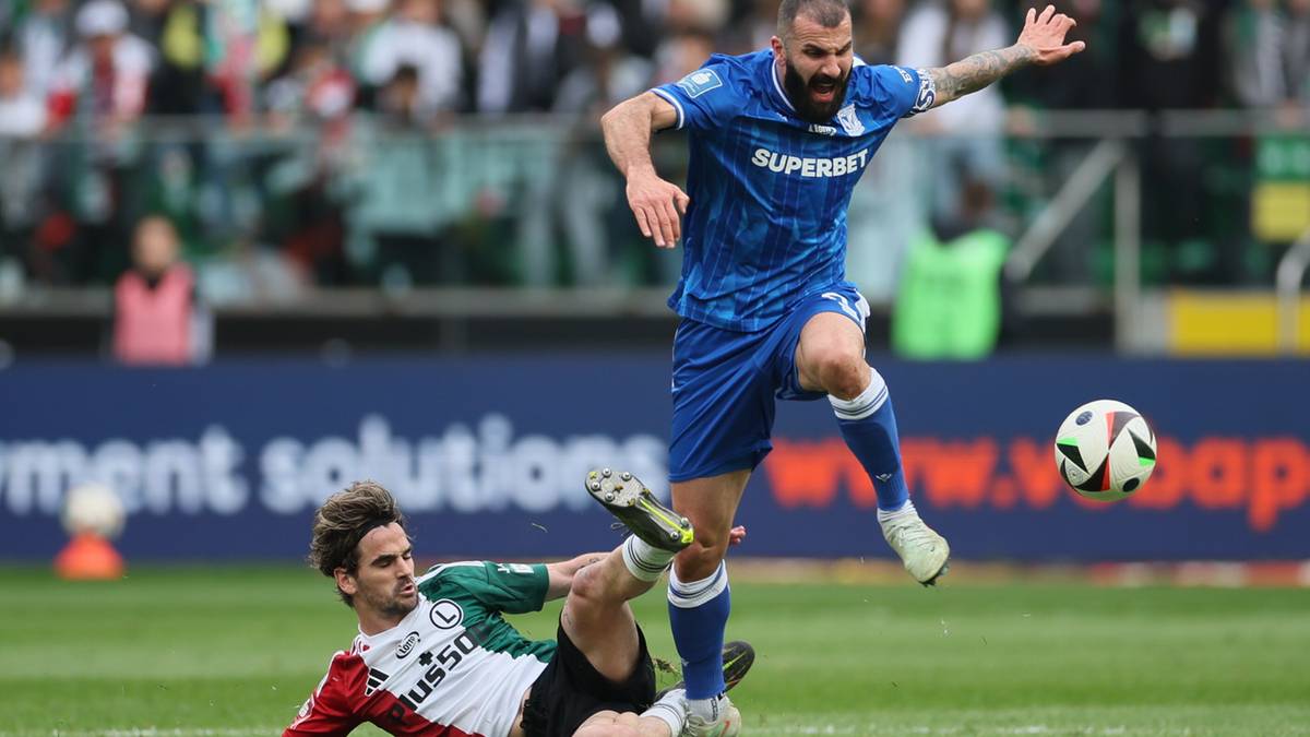 Dwóch piłkarzy walczących o piłkę na boisku, jeden w niebieskiej koszulce próbuje obronić się przed przeciwnikiem w zielono-białej koszulce, który leży na ziemi.