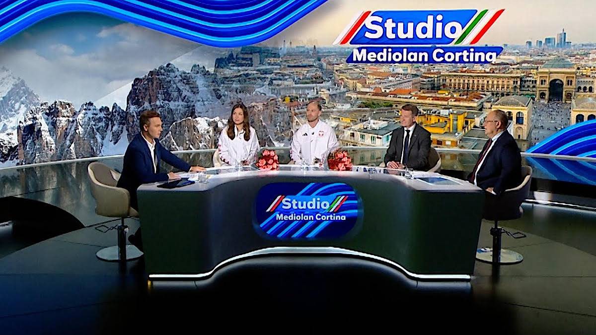 Cztery osoby siedzą przy stole w studiu telewizyjnym, w tle widok na miasto i góry, nad stołem logo "Studio Mediolan Cortina".